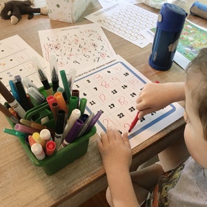 May include: A child is working on a number puzzle, filling in the missing numbers on a grid. The child is using a red marker to write the numbers. There are many colored markers and pens in a green container.