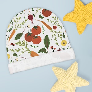 Puede incluir: Un gorro de bebé blanco con un colorido estampado de verduras, que incluye tomates, zanahorias y remolachas. El gorro tiene un borde enrollado y está decorado con hojas verdes y pequeñas flores. Dos juguetes amarillos en forma de estrella están en el fondo.