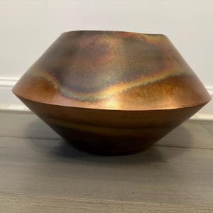 Puede incluir: Un jarrón grande de metal de color cobre con una abertura ancha y acampanada y un fondo redondeado. El jarrón tiene un acabado desgastado y envejecido con tonos de marrón, naranja y verde.