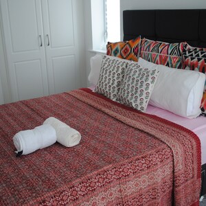 May include: A red and white patterned bedspread with two white towels on top. The bed has a pink sheet and a black headboard with colourful patterned pillows.