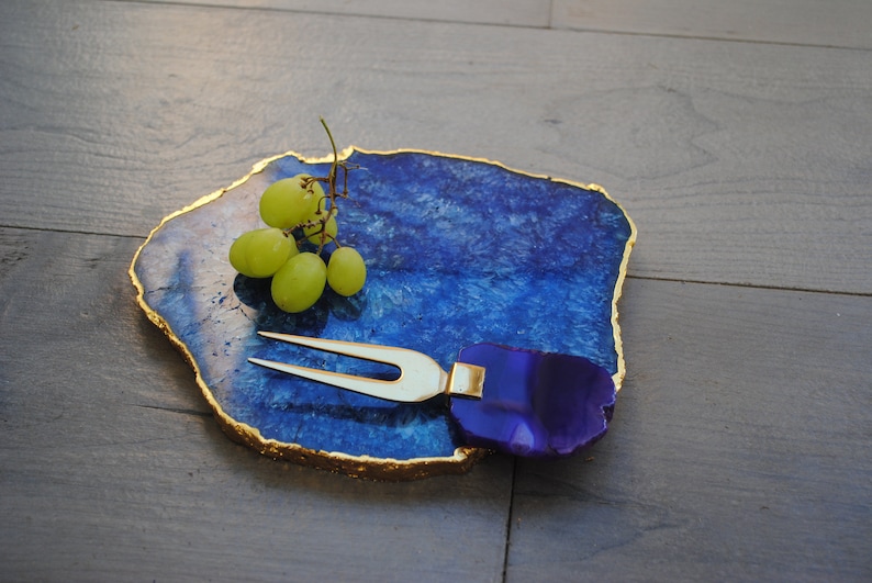 Puede incluir: Una tabla de servir de &aacute;gata azul con bordes dorados, combinada con un tenedor para queso. Un racimo de uvas verdes descansa sobre la tabla. La forma irregular y los patrones naturales de la tabla crean una est&eacute;tica &uacute;nica.