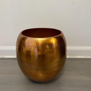 Puede incluir: Un macetero grande de forma ovalada de color cobre con una superficie ligeramente texturizada.