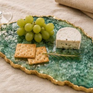 Puede incluir: Una bandeja decorativa con borde verde y dorado, con uvas verdes, galletas y un trozo de queso. La bandeja tiene un borde dorado y una apariencia texturizada similar a una gema.