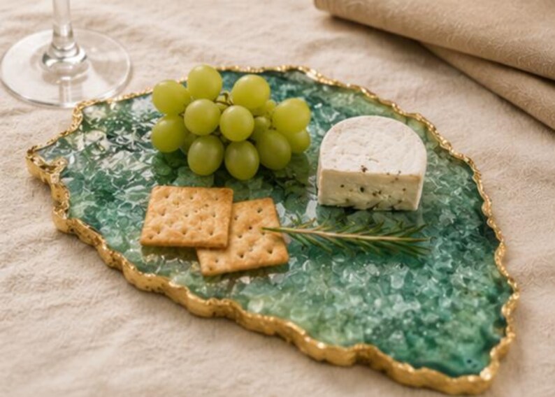 Puede incluir: Una bandeja decorativa con borde verde y dorado, con uvas verdes, galletas y un trozo de queso. La bandeja tiene un borde dorado y una apariencia texturizada similar a una gema.