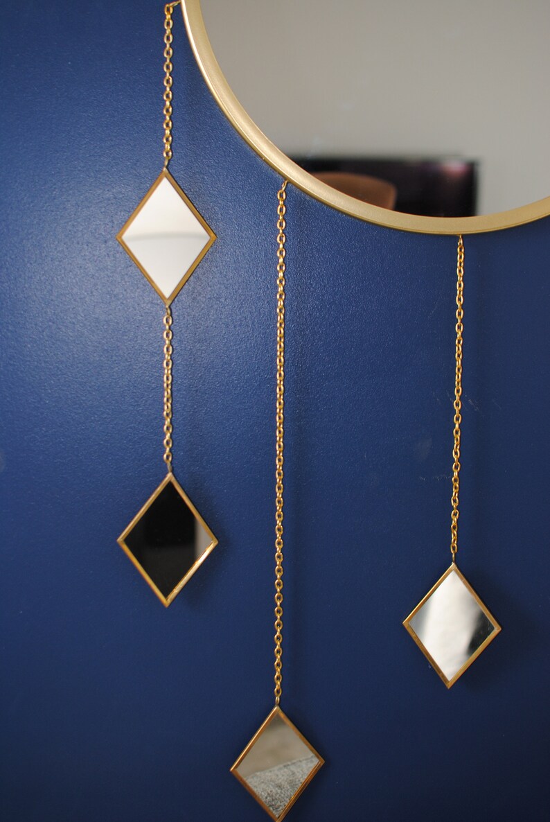 May include: A round mirror with a gold frame and three smaller diamond-shaped mirrors hanging from chains. The smaller mirrors are black, white, and silver.