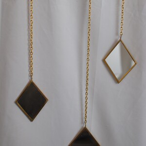 May include: Three gold-toned diamond-shaped mirrors hang from gold chains against a white background. The mirrors are different sizes and are all facing forward.