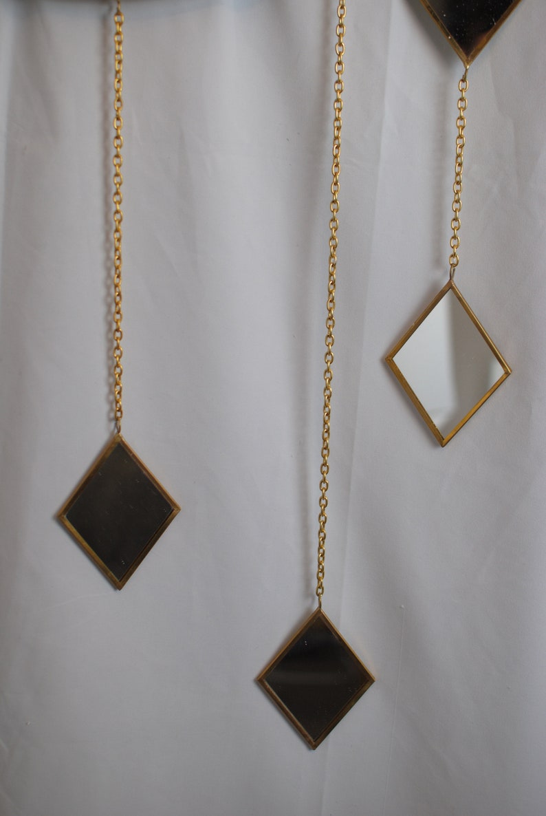 May include: Three gold-toned diamond-shaped mirrors hang from gold chains against a white background. The mirrors are different sizes and are all facing forward.