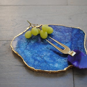 Puede incluir: Una tabla de servir de &aacute;gata azul con bordes dorados, que sostiene un racimo de uvas verdes. Un tenedor para queso dorado con un mango morado descansa sobre la tabla. La tabla est&aacute; sobre una superficie de madera oscura.