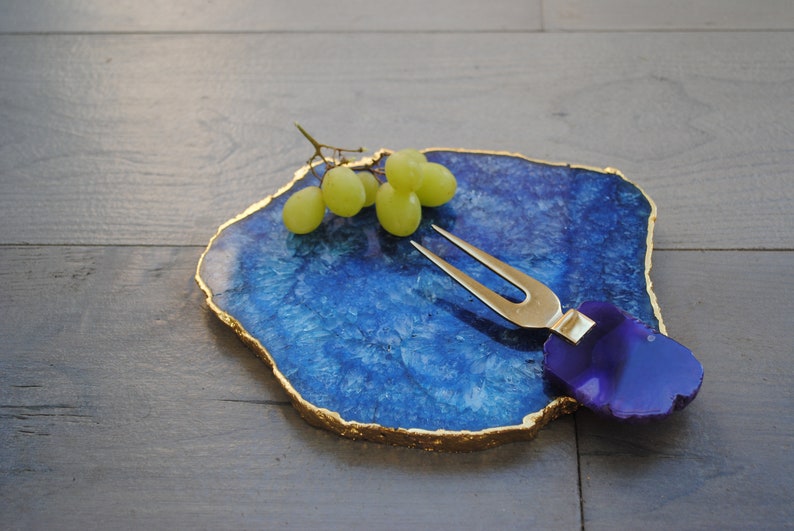 Puede incluir: Una tabla de servir de &aacute;gata azul con bordes dorados, que sostiene un racimo de uvas verdes. Un tenedor para queso dorado con un mango morado descansa sobre la tabla. La tabla est&aacute; sobre una superficie de madera oscura.