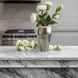 May include: A silver hammered metal vase with white roses on a white marble countertop.