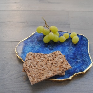 Puede incluir: Una bandeja de servicio de &aacute;gata azul con borde dorado, con uvas verdes y dos galletas. La bandeja tiene un acabado pulido, ideal para servir aperitivos. Dimensiones: aproximadamente 25 cm.