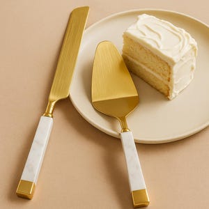 Op de afbeelding: Een gouden taartmes en -serveerset met witte marmeren handvatten en gouden accenten. Een stuk cake met witte glazuur ligt op een crèmekleurige plaat. Voor het snijden en serveren van taart.