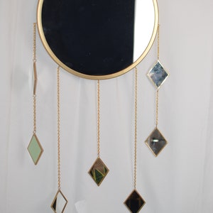 May include: A round gold-framed mirror with a black reflection hangs on a white wall. Five smaller diamond-shaped mirrors hang from gold chains attached to the larger mirror.