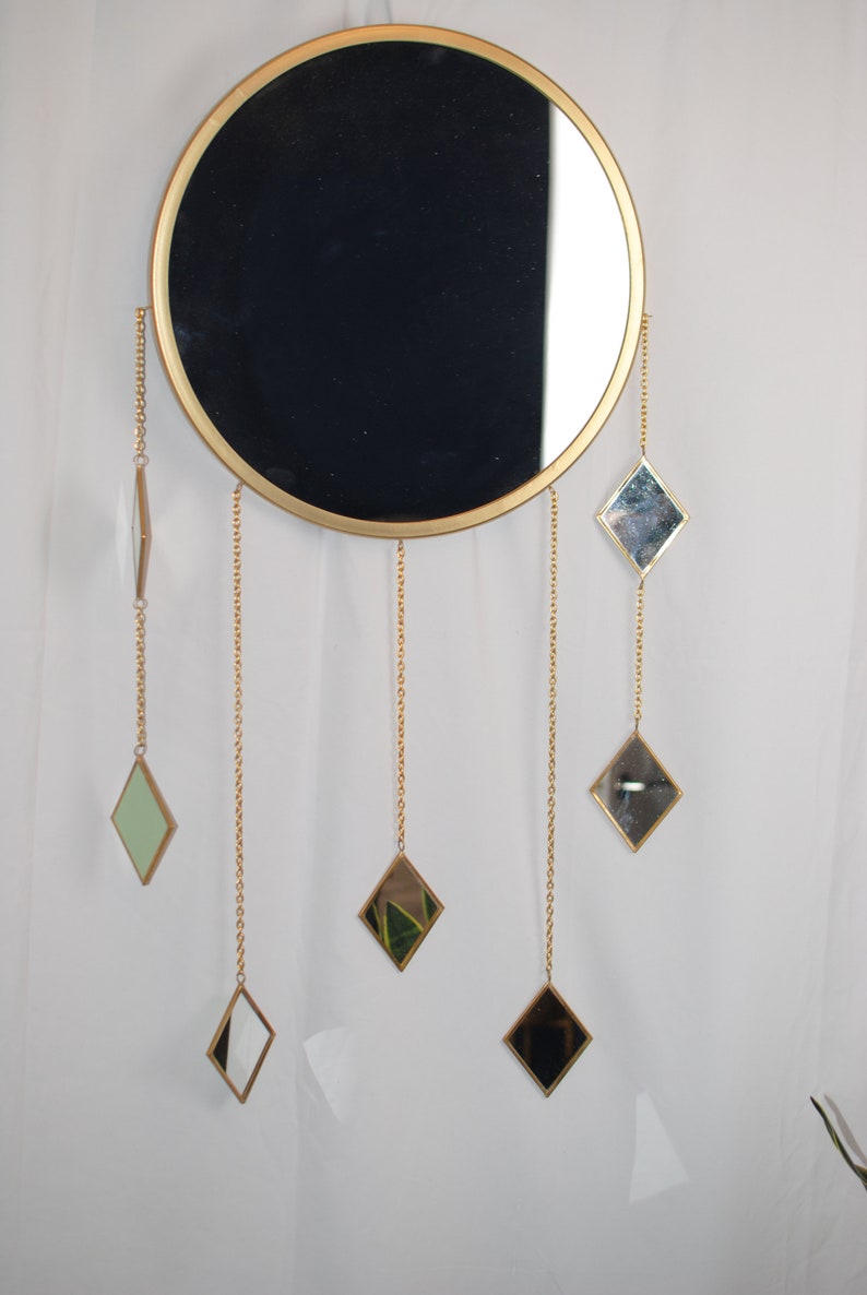 May include: A round gold-framed mirror with a black reflection hangs on a white wall. Five smaller diamond-shaped mirrors hang from gold chains attached to the larger mirror.