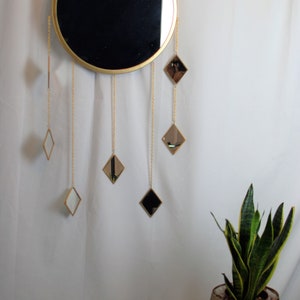 May include: A gold round mirror with a black reflective surface hangs on a white wall. Five smaller diamond-shaped mirrors hang from gold chains below the larger mirror.
