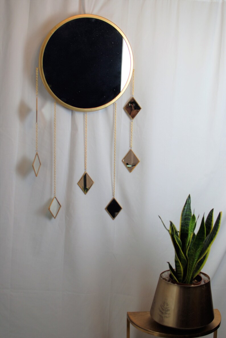 May include: A gold round mirror with a black reflective surface hangs on a white wall. Five smaller diamond-shaped mirrors hang from gold chains below the larger mirror.