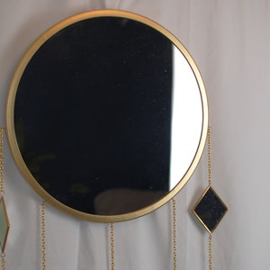 May include: A round gold-framed mirror with three dangling chains and diamond-shaped mirrors at the ends. The mirror is hanging on a white wall.