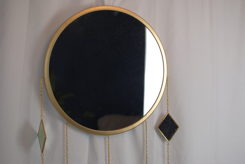 May include: A round gold-framed mirror with three dangling chains and diamond-shaped mirrors at the ends. The mirror is hanging on a white wall.