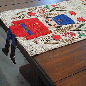 May include: A white beaded table runner with a red, blue, and green embroidered nutcracker design. The nutcracker has a blue hat and red and white uniform. The runner is on a dark brown wooden table.