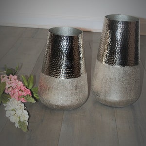 May include: Two silver vases with a hammered texture on the top half and a frosted appearance on the bottom half. The vases are displayed on a wooden surface, with a bouquet of pink and white flowers to the left.