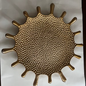 K&ouml;nnte beinhalten: Ein goldfarbenes Tablett aus Metall mit einer strukturierten Oberfl&auml;che in Form einer stilisierten Sonnenstrahlen oder eines Spritzers von Fl&uuml;ssigkeit. Das Tablett hat eine kreisf&ouml;rmige Mitte und 12 nach au&szlig;en ragende Punkte.