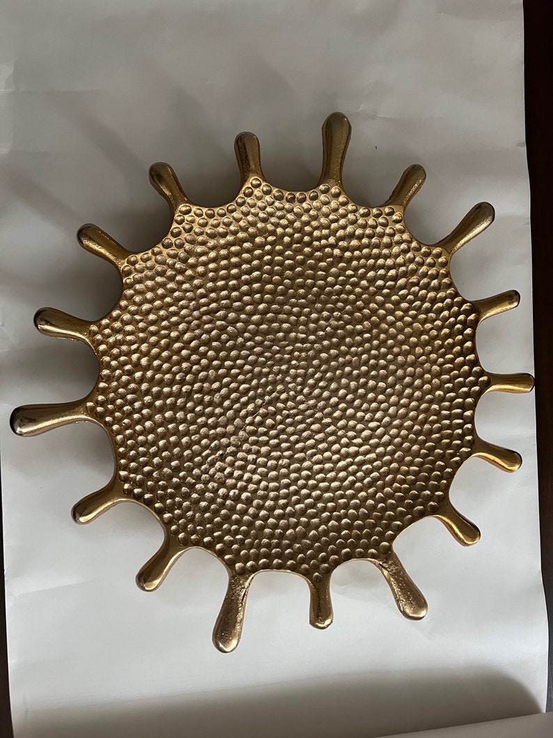 K&ouml;nnte beinhalten: Ein goldfarbenes Tablett aus Metall mit einer strukturierten Oberfl&auml;che in Form einer stilisierten Sonnenstrahlen oder eines Spritzers von Fl&uuml;ssigkeit. Das Tablett hat eine kreisf&ouml;rmige Mitte und 12 nach au&szlig;en ragende Punkte.