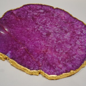 Puede incluir: Una rebanada decorativa de &aacute;gata p&uacute;rpura con un borde dorado. El &aacute;gata tiene una estructura cristalina y una superficie pulida. El borde dorado a&ntilde;ade un toque de elegancia a esta piedra natural.