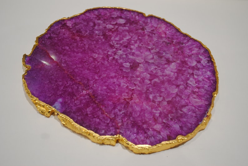 Puede incluir: Una rebanada decorativa de &aacute;gata p&uacute;rpura con un borde dorado. El &aacute;gata tiene una estructura cristalina y una superficie pulida. El borde dorado a&ntilde;ade un toque de elegancia a esta piedra natural.