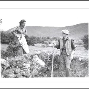 Peut inclure: Une photographie en noir et blanc montrant deux personnages dans un paysage rural. L'un porte une chemise à manches longues et un tablier, tandis que l'autre porte une chemise à col, un gilet, un pantalon et une casquette, tenant un outil à long manche.
