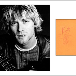 May include: A black and white portrait of Kurt Cobain with blonde hair, wearing a dark jacket over a patterned shirt, holding a guitar neck. Beside the portrait is a rectangular orange card featuring the handwritten signature Kurt Cobain.
