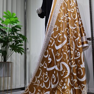 May include: A brown satin scarf with a white geometric pattern and Arabic calligraphy. The scarf is draped over a mannequin.