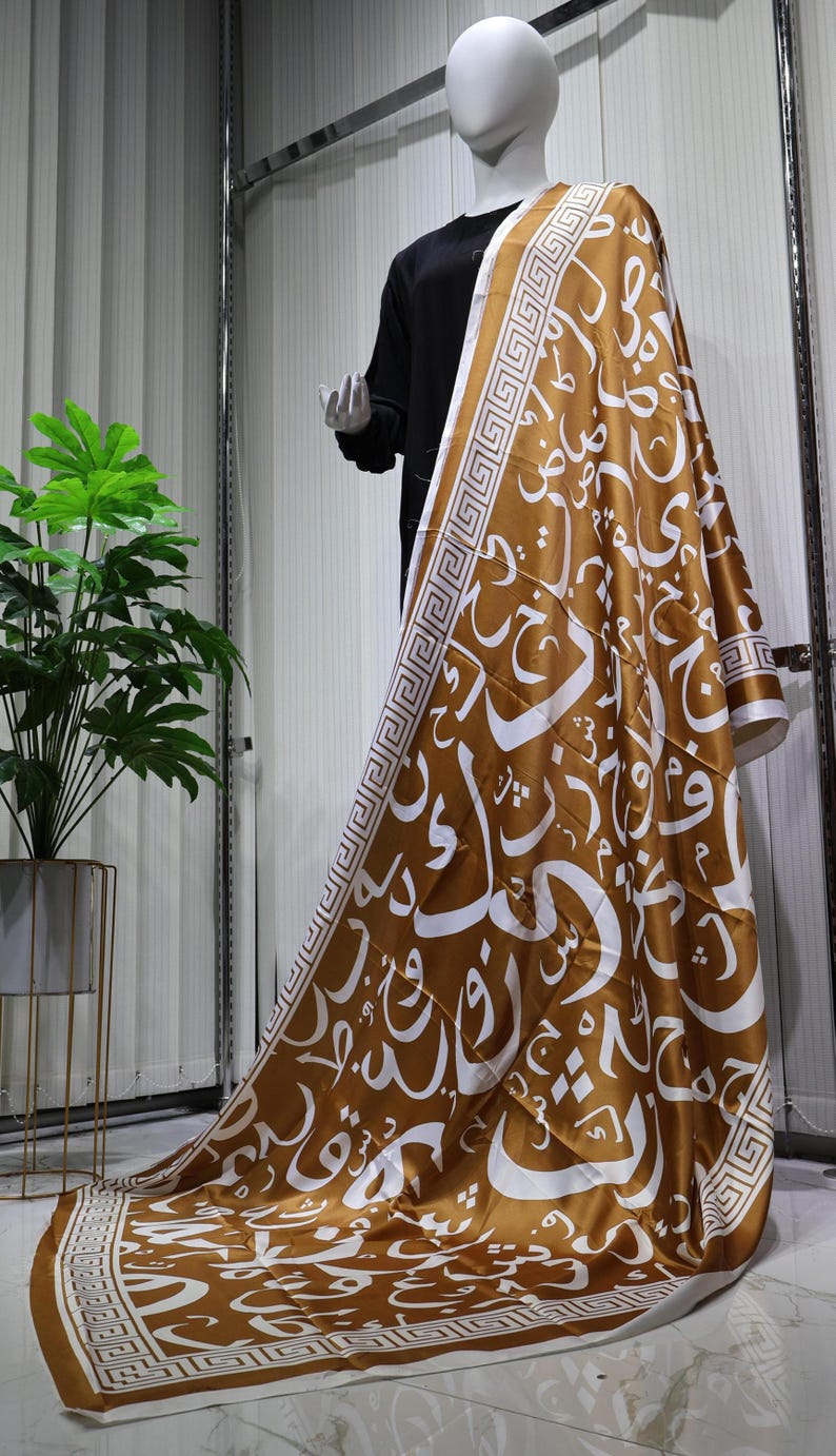 May include: A brown satin scarf with a white geometric pattern and Arabic calligraphy. The scarf is draped over a mannequin.