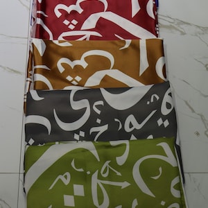 May include: A stack of six fabric squares with white Arabic calligraphy on a variety of coloured backgrounds, including blue, red, gold, grey, and green.
