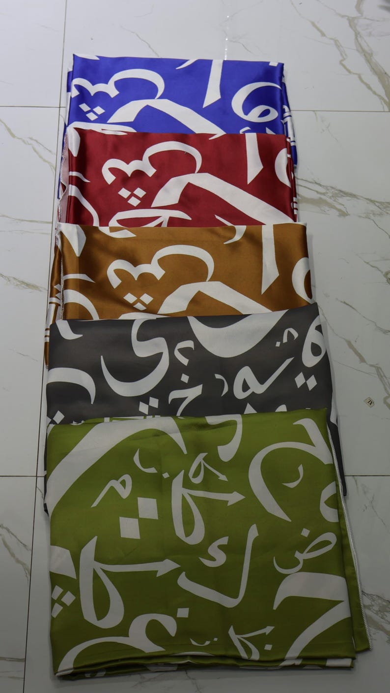 May include: A stack of six fabric squares with white Arabic calligraphy on a variety of coloured backgrounds, including blue, red, gold, grey, and green.