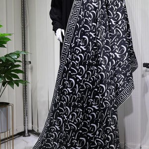 May include: A black and white patterned scarf with a geometric border. The scarf features a design of Arabic calligraphy.