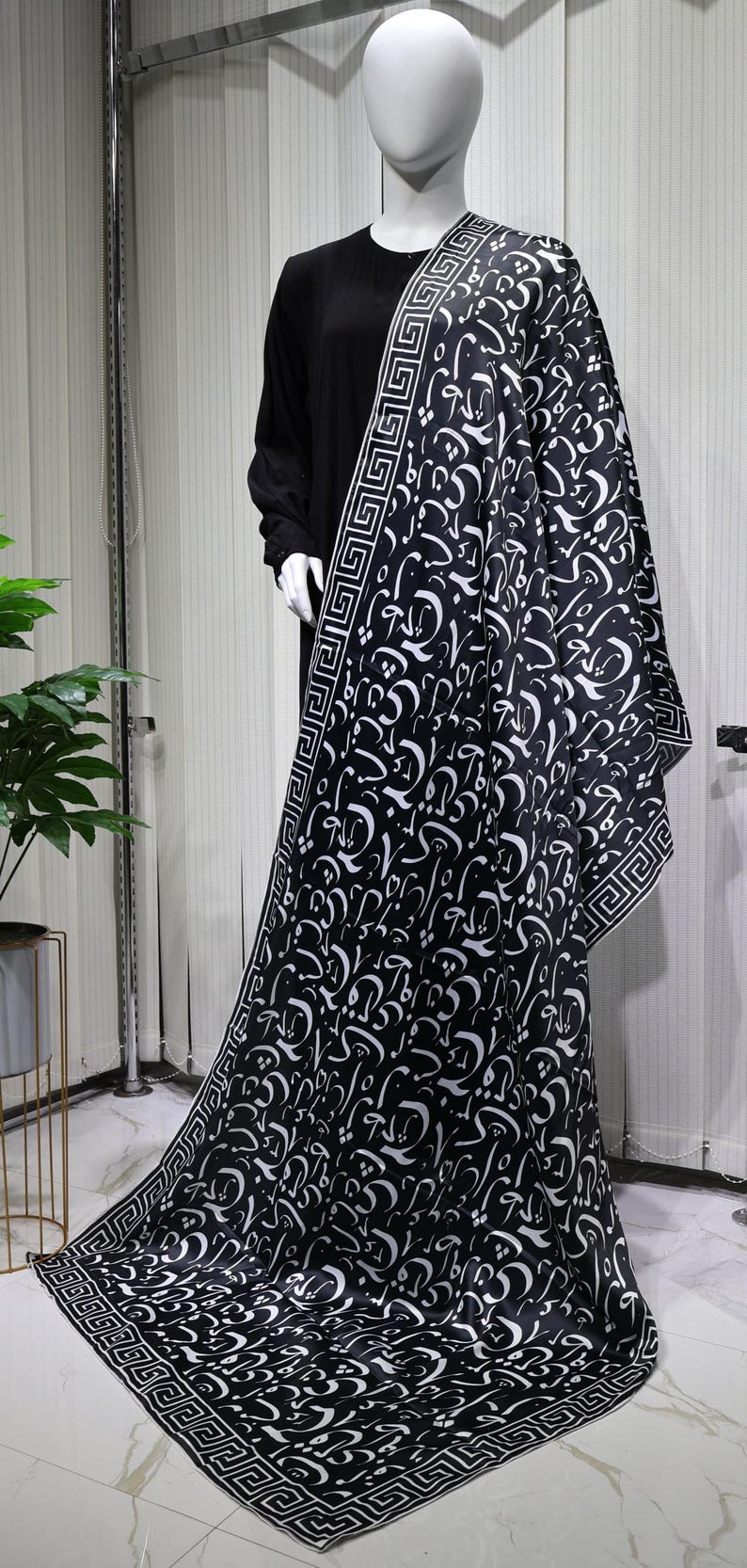 May include: A black and white patterned scarf with a geometric border. The scarf features a design of Arabic calligraphy.