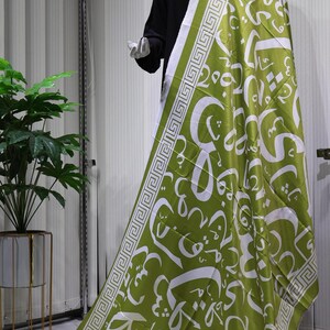 May include: A green and white patterned scarf with a geometric border. The scarf features Arabic calligraphy in white on a green background.