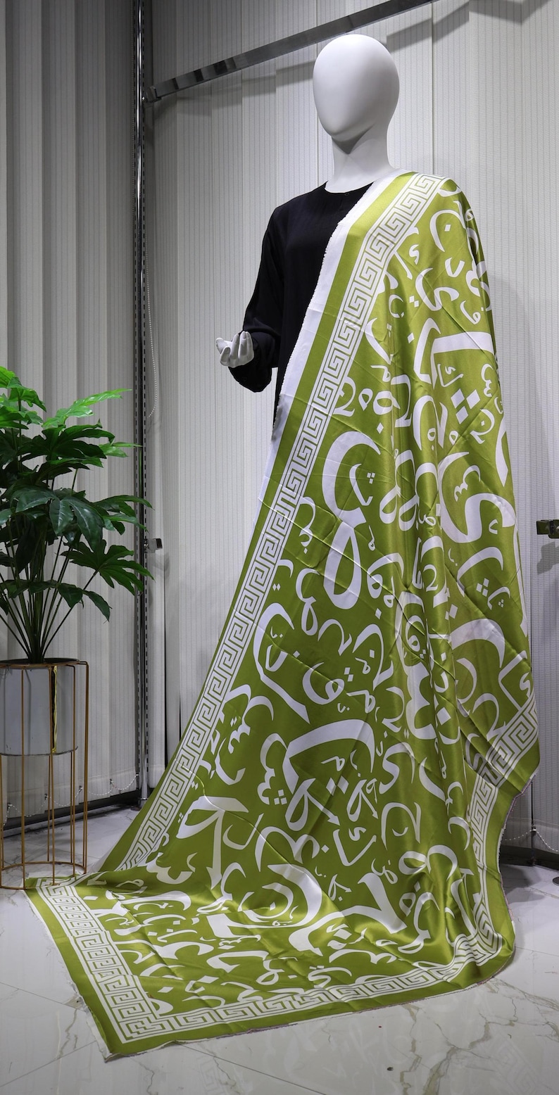 May include: A green and white patterned scarf with a geometric border. The scarf features Arabic calligraphy in white on a green background.