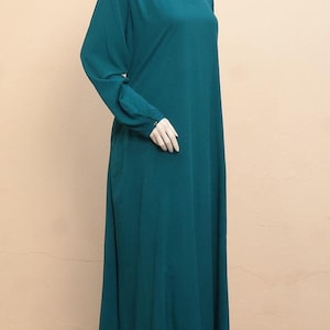 May include: A long-sleeved, floor-length dress in a solid deep teal color. The garment features a simple round neckline with a small front slit detail and long sleeves that end in buttoned cuffs. The silhouette is loose and flows to the floor.