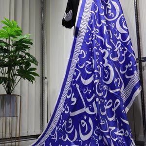 May include: A blue and white scarf featuring a geometric pattern and Arabic calligraphy. The scarf is made from a silky material and has a fringed edge.