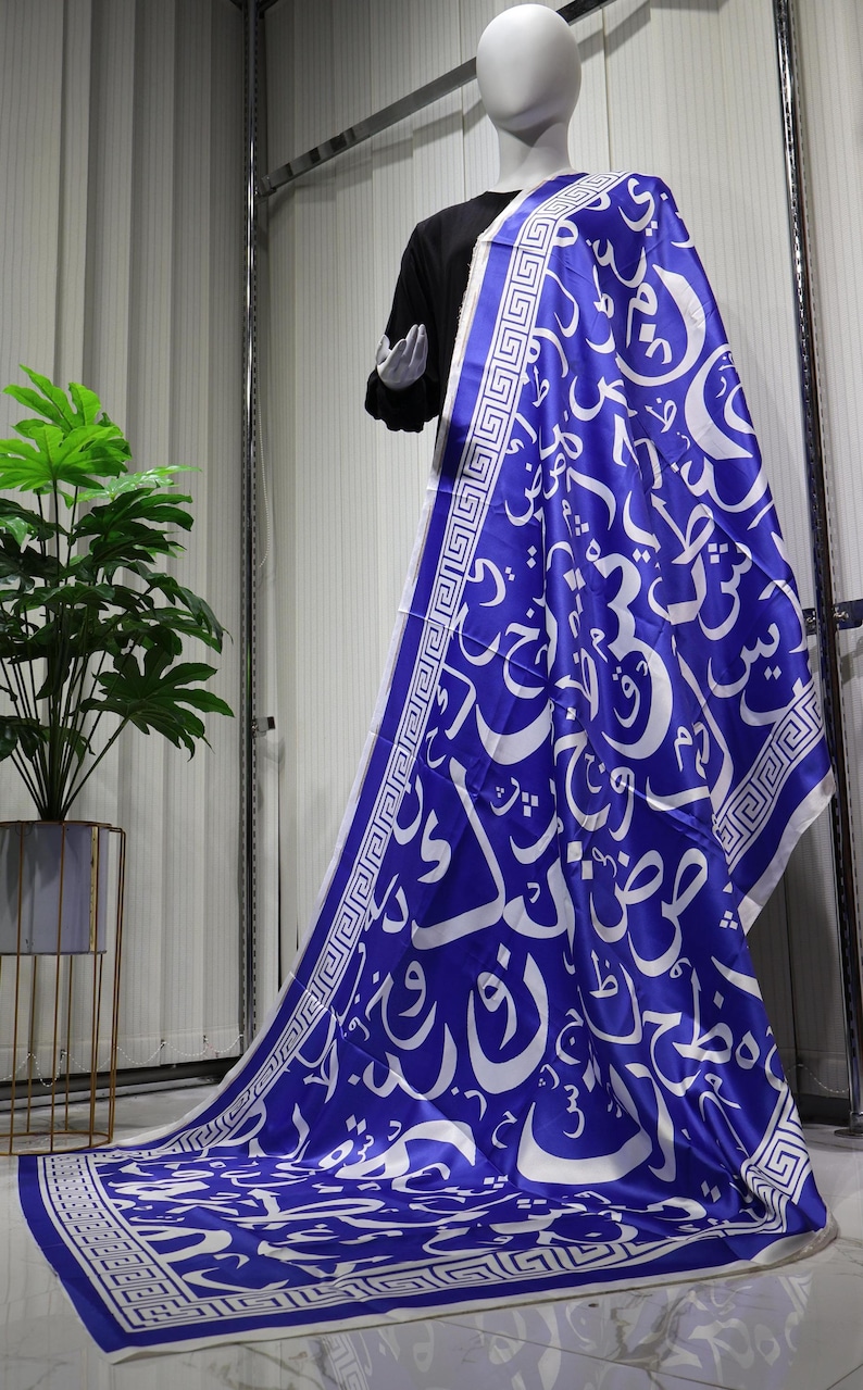 May include: A blue and white scarf featuring a geometric pattern and Arabic calligraphy. The scarf is made from a silky material and has a fringed edge.