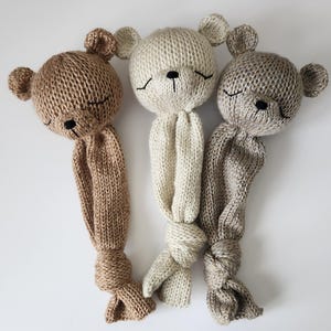 May include: Three knitted teddy bear toys with closed eyes. The bears are beige, brown, and light brown. The bears are tied in a knot at the bottom.