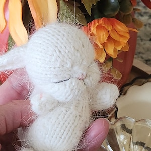 May include: A white knitted bunny with closed eyes, sitting in a hand. The bunny is soft and fluffy, with a delicate knitted texture.