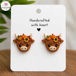 May include: A pair of stud earrings featuring brown Highland cow heads adorned with orange pumpkins, yellow sunflowers, and red autumn leaves. The earrings are displayed on a white card with the text Handcrafted with heart and a black heart icon.