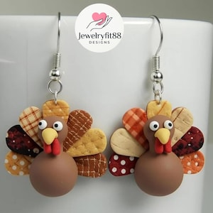May include: A pair of handmade turkey earrings. Each earring features a brown turkey body, a yellow beak, and a red wattle. The turkey's tail feathers are made of various patterned fabrics in shades of brown, orange, and cream. The earrings hang from silver-colored hooks.