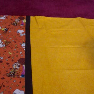 Könnte beinhalten: Ein Stoffpanel mit schwarzem Rand und einem bunten Druck mit Comicfiguren. Der Druck ist auf orangefarbenem Grund mit gelben, braunen und weißen Details. Die andere Seite des Panels ist einfarbig gelb.