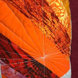 Puede incluir: Primer plano de una almohada acolchada con un patr&oacute;n geom&eacute;trico de tela naranja, roja y marr&oacute;n. La almohada est&aacute; cosida con hilo blanco en un patr&oacute;n radiante.