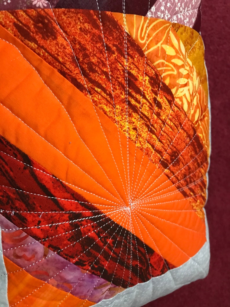 Puede incluir: Primer plano de una almohada acolchada con un patr&oacute;n geom&eacute;trico de tela naranja, roja y marr&oacute;n. La almohada est&aacute; cosida con hilo blanco en un patr&oacute;n radiante.