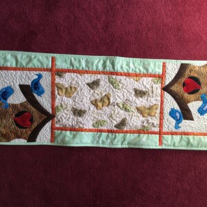 May include: A green and brown quilted table runner with a patchwork design featuring butterflies, birds, and hearts. The words "home" are embroidered on each end of the runner.