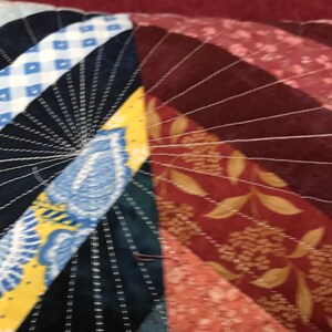 Puede incluir: Primer plano de una colcha con una variedad de telas y colores, incluyendo azul, amarillo, rojo y blanco. La colcha est&aacute; cosida en un patr&oacute;n radial, con las l&iacute;neas de costura que irradian desde el centro.